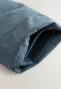 Blaues, glattes, mattes Außenmaterial gibt teilweise einen Blick auf das dunkle, gerippte Innenfutter frei. Die Kanten sind sauber mit strukturellen Nähten genäht.