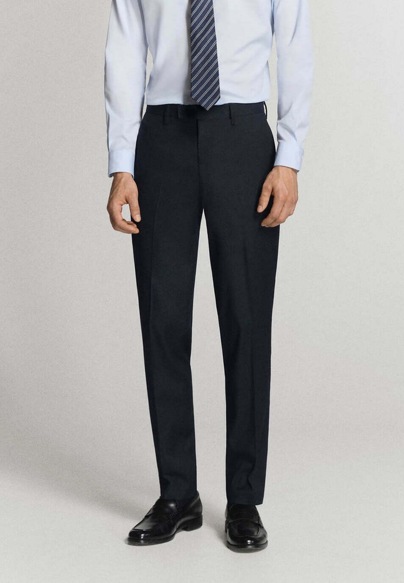 Homme portant un pantalon formel noir, une chemise bleu clair, une cravate rayée et des chaussures habillées noires, debout sur un fond uni.