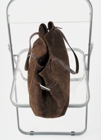 Bolso tote de ante marrón con doble asa, colocado sobre una silla plegable transparente contra un fondo blanco.