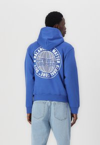 Blaue Sweatshirt mit Kapuze, mit einem großen weißen Grafikdesign auf dem Rücken und gerippten Bündchen und Saum. Kombiniert mit hellblauen Jeans.