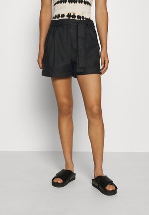 Femme portant un short noir ceinturé et des sandales noires, debout devant un fond clair uni.