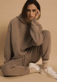Jasnobrązowy, oversize'owy hoodie i dresy z wytłaczanym napisem, w połączeniu z białymi sneakersami. Miękka tkanina i luźny krój. Neutralne tło.