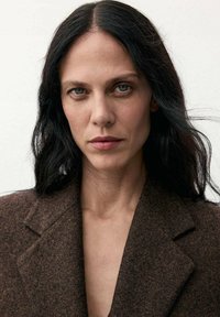 Una persona che indossa un blazer di lana marrone con colletto a punte, caratterizzato da un tessuto testurizzato e una leggera lucentezza, su uno sfondo semplice.