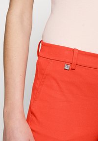 Nära bild av en person som bär röda högmidjade byxor med bälteshål och en silverfyrkantig detalj nära midjan.