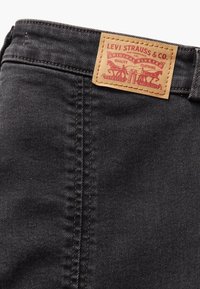 Jeans de mezclilla negra con un parche de cuero marrón claro con un logo en relieve rojo, diseño remachado y costura en contraste en la cinturilla.