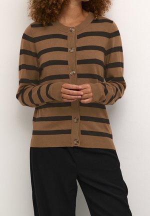 Vrouw draagt een bruine cardigan met zwarte horizontale strepen en een zwarte broek, staand tegen een effen witte achtergrond.