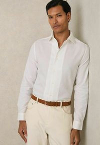 Cască albă cu mânecă lungă și guler cu nasturi, asociată cu pantaloni de culoare deschisă și o curea din piele maro. Textură netedă, croi ajustat.