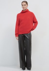 Eine Frau steht da und trägt einen roten Rollkragenpullover, schwarze breite Lederhosen, schwarze Schuhe und silberne Creolen vor einem weißen Hintergrund.
