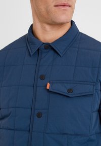 Navy quilted shirt jacket with a button-up front, black buttons, and a chest pocket featuring an orange accent. Smooth, lightweight fabric.