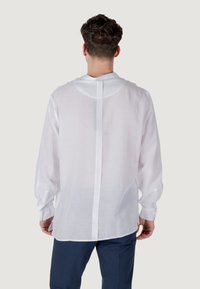 Camicia bianca a maniche lunghe realizzata in tessuto leggero, con scollo rotondo, cucitura centrale sul retro e polsini con bottoni. Vestibilità tailored.