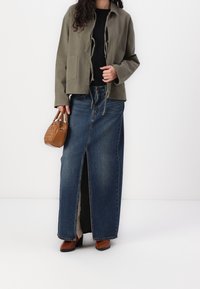 Vihreä oliivinvihreä takki naruilla, musta paita ja korkeavyötäröinen denim-maksihame, jossa on edessä halkio. Ruskea käsilaukku ja korkeat ruskeat saappaat.