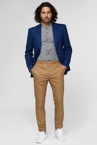 Blazer blu a righe sottili sopra una camicia grigia con bottoni, abbinato a pantaloni beige e scarpe da ginnastica bianche. Tessuto liscio, vestibilità su misura.