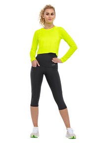 Neon keltainen pitkähihainen crop-top, joka on yhdistetty mustiin capri-legginseihin; topissa on logo, leggingseissä on sivutaskut ja ne ovat istuvat.