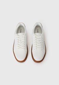 Een paar witte leren sneakers met bruine zolen, witte veters en Calvin Klein branding aan de binnenkant en op de tong, gezien van bovenaf.