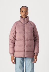 Pastellrosa polstret jakke med høy krage, lange ermer, ribbestrikkede mansjetter og horisontale sømdetaljer. Kombinert med lyseblå denimbukser.