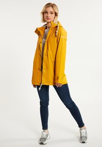 Chaqueta de lluvia amarilla impermeable con capucha, cordón ajustable, botones a presión y bolsillos laterales. Combinada con jeans oscuros y zapatillas gris.