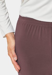 Bordeaux kleurige stretchbroek met een elastische tailleband, een gladde textuur en een aansluitende pasvorm, gedeeltelijk getoond naast een hand die op de stof ligt.