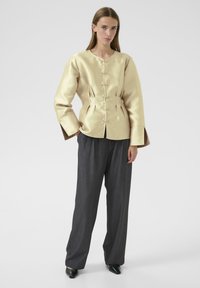 Satin gold jacket with a high neckline, front buttons, and pleats, paired with gray wide-leg trousers and black pointed shoes.