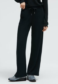 Schwarze, gerippte, breite Hose mit elastischem Bund und Kordelzug. Kombiniert mit grauen Sportschuhen mit blauen Akzenten.