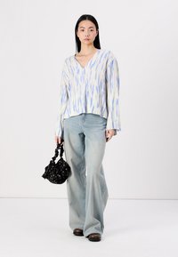 Hellbluse mit einem Muster in Blau und Weiß, V-Ausschnitt, langen Ärmeln und Knöpfen, kombiniert mit hellblauen, ausgestellten Jeans und einer schwarzen Tasche.