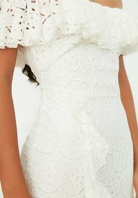 Valkoinen olkaimeton mekko, jossa on kukkakuvioista pitsiä, röyhelöreunus, kapeampi vyötärö ja ympyräkirjontakuviot koko kankaan alueella.