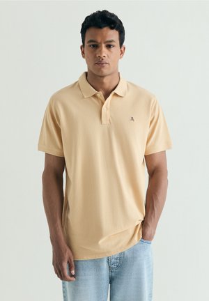 Joven con cabello rizado oscuro, vistiendo una camiseta polo beige clara y vaqueros azul claro sobre un fondo liso.