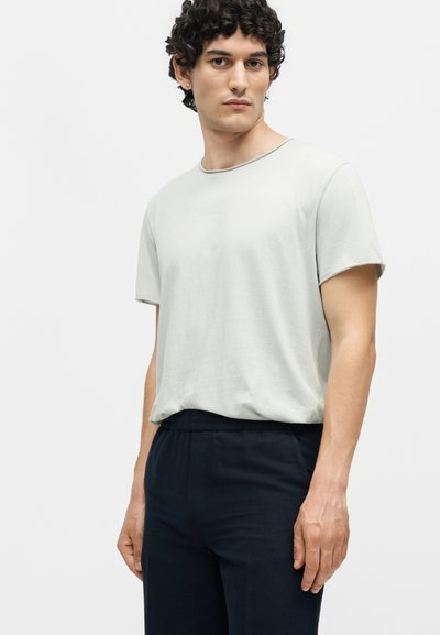 Νέος άνδρας με σγουρά μαλλιά που φοράει ανοιχτό γκρι κοντομάνικη μπλούζα με στρογγυλή λαιμόκοψη και σκούρο μπλε παντελόνι σε λευκό, μονόχρωμο φόντο.