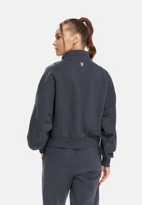 Gekürzter grauer Sweatshirt mit hohem Kragen, strukturiertem Stoff und kleinem weißen Logo auf dem Rücken, breiten Ärmeln und einem taillierten Bund.