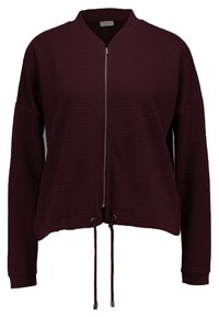 Viininpunainen lyhyt takki, jossa on ribbitekstuuri, vetoketjulausunto, sivutaskut ja säädettävä nyöri helman kohdalla. Varustettu pystykauluksella.