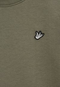 Olivgrön sweatshirt med en texturerad tyg, rund halsringning och en liten vit fågel broderad på vänster sida.