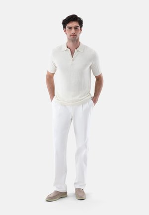 Mann steht und trägt ein weißes kurzärmeliges, strukturiertes Poloshirt, weiße Hose, beige Slipper, Hände in den Taschen, neutraler Hintergrund.