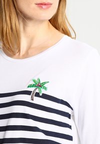 Camicia a righe bianche con un distintivo ricamato di una palma verde e rosa nella parte sinistra del petto. Tessuto in cotone morbido e vestibilità rilassata.