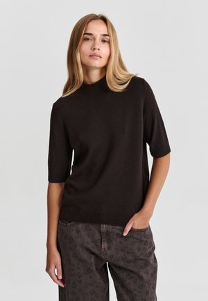 Jeune femme aux longs cheveux blonds portant un pull noir à manches courtes et un pantalon foncé à motifs, debout devant un fond uni.