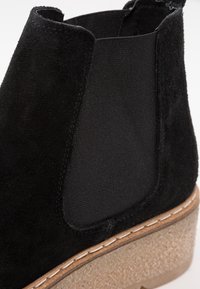 Bottine Chelsea en daim noir avec panneau latéral élastique et semelle en crêpe beige texturée, vue en gros plan.