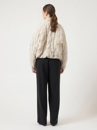 Béžová prošívaná bunda s vysokým límcem a texturovaným vzorem, doplněná černými širokými kalhotami. Pohled zezadu.