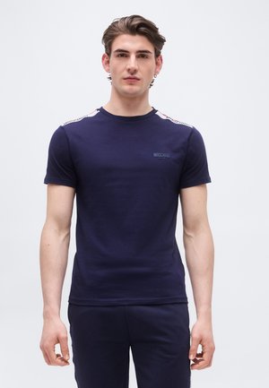 Jeune homme aux cheveux bruns courts portant un t-shirt bleu marine Moschino Active et un pantalon assorti, debout devant un fond blanc uni.