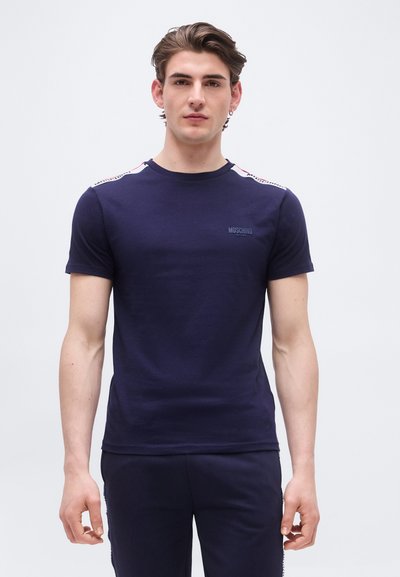 Jeune homme aux cheveux bruns courts portant un t-shirt bleu marine Moschino Active et un pantalon assorti, debout devant un fond blanc uni.