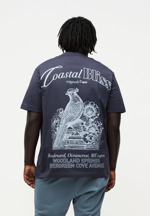 Personne aux dreadlocks foncés porte une chemise bleu marine avec une grande illustration blanche d'oiseau, des fleurs et un texte incluant "Coastal Bliss" et les détails de localisation.