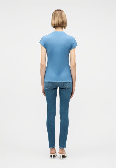 Άτομο με κοντά ξανθά μαλλιά, ντυμένο με εφαρμοστή μπλε κοντομάνικη μπλούζα, μπλε στενά τζιν και λευκά παπούτσια με τακούνι, όρθιο πλάτη προς τον θεατή.