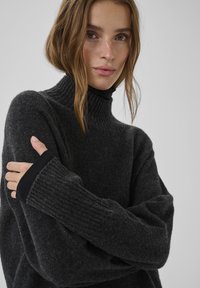 Tumši pelēks džemperis ar ribotu augsto apkakli un aprocēm. Mīksta tekstūra, brīvs piegriezums un garas piedurknes ar melniem akcentiem aprocēs.