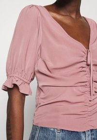 Blusa rosa con escote en V, mangas abullonadas y detalles fruncidos. Confeccionada en material suave, presenta un cierre con lazo en la parte delantera.