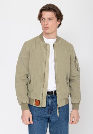 Jonge man met een kaki bomberjack over een wit overhemd en blauwe spijkerbroek, staande tegen een effen lichte achtergrond en naar links kijkend.