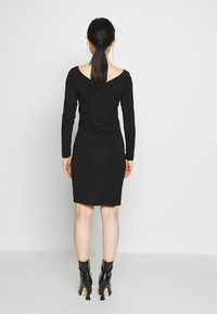 Svart långärmad klänning med off-shoulder-design, åtsittande form och ruchingdetaljer på baksidan, kombinerad med svarta ankelskor.