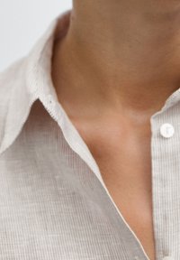 Jasno beżowa lniana koszula z wyrazistym kołnierzykiem, z delikatną prążkowaną teksturą i pojedynczym guzikiem przy dekolcie.