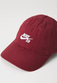 Caciula de baseball burgundy cu umbră curbă, realizată din bumbac. Prezintă un logo brodat alb "Nike SB" pe față. Design simplu cu detalii de cusătură.
