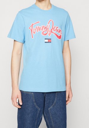 Jasnoniebieska bawełniana koszulka z wyraźnym czerwonym nadrukiem "Tommy Jeans" oraz małą naszywką z logo w kolorach granatowym i czerwonym na przodzie.