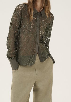 Femme portant une chemise boutonnée en dentelle florale vert foncé avec col et poignets unis, associée à un pantalon beige taille haute.