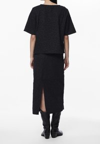 Woman wearing a black textured short-sleeve top and matching midi skirt with a back slit, paired with black mid-calf boots, standing against white background.