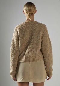 Beige Strickpullover mit einer strukturierten, flauschigen Oberfläche, überschnittenen Schultern und gerippten Bündchen. Kombiniert mit einem kurzen, hellen Rock.