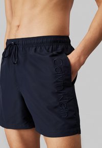 Shorts de bain navy avec une taille élastique et un cordon de serrage. Comprend des poches latérales et le logo "Calvin Klein" brodé en fil sombre.